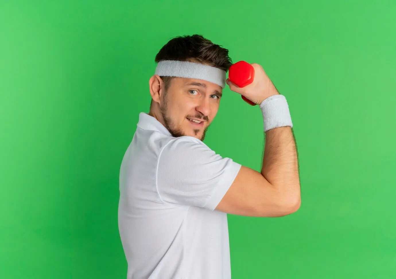 Молодой мужчина, занимающийся фитнесом, в белой футболке и с повязкой на голове, напряжённый и уверенный, тренируется с гантелью на фоне зелёной стены.